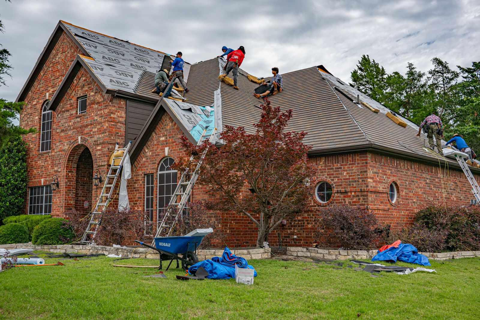 Roofing crew replacing asphalt shingles on a brick suburban home, ladders and underlayment visible
