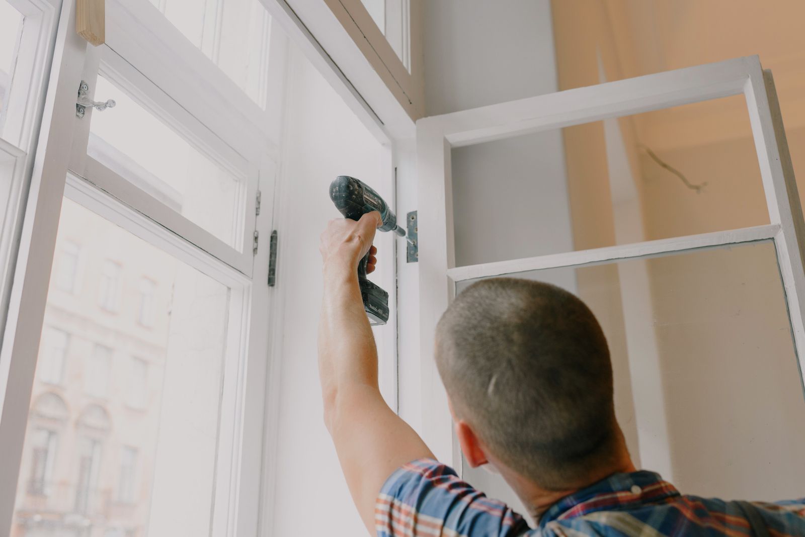 Installer seen from behind assembling a white-framed window with a cordless drill